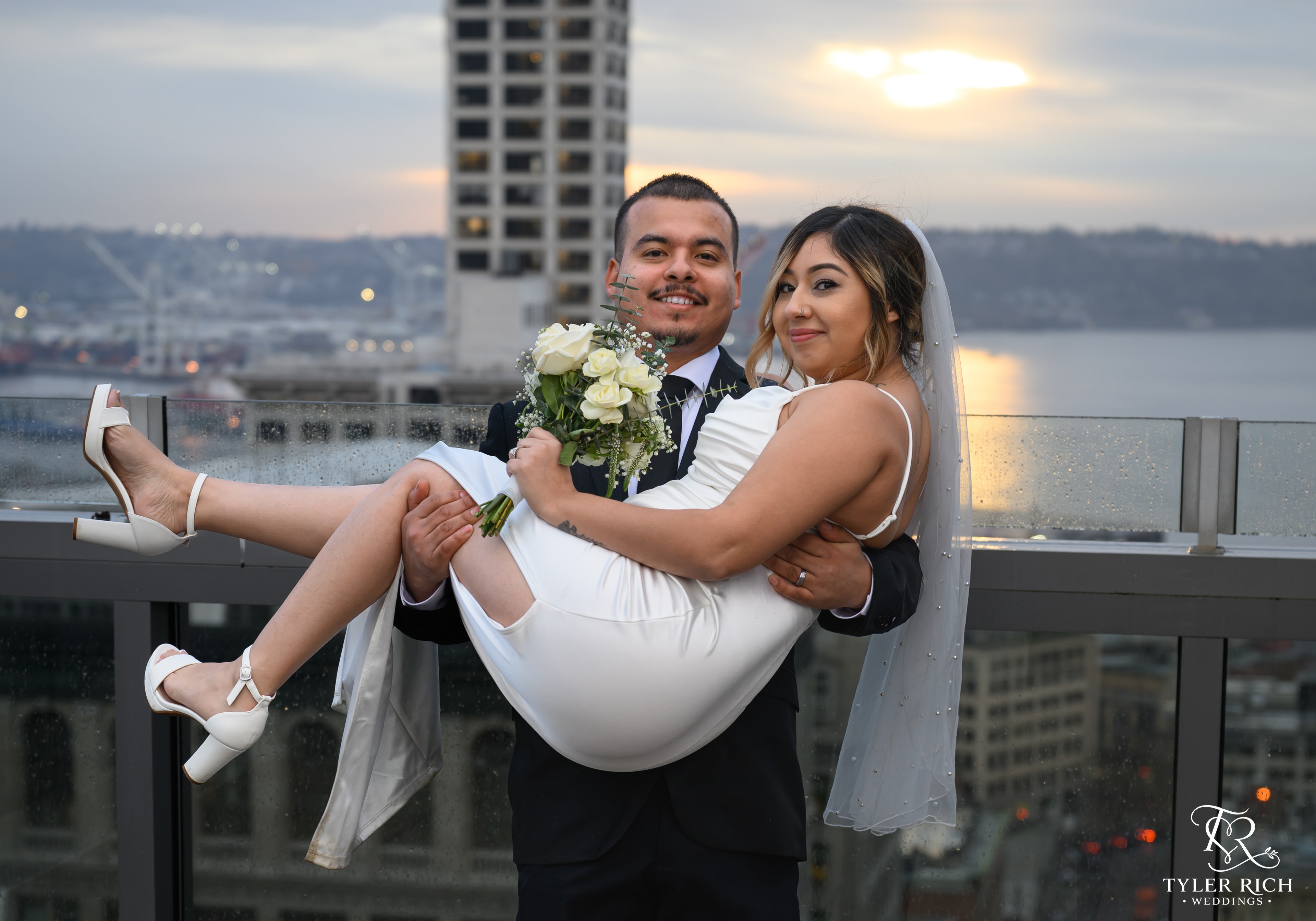 Groom carrying bride on a rooftop with city skyline at sunset